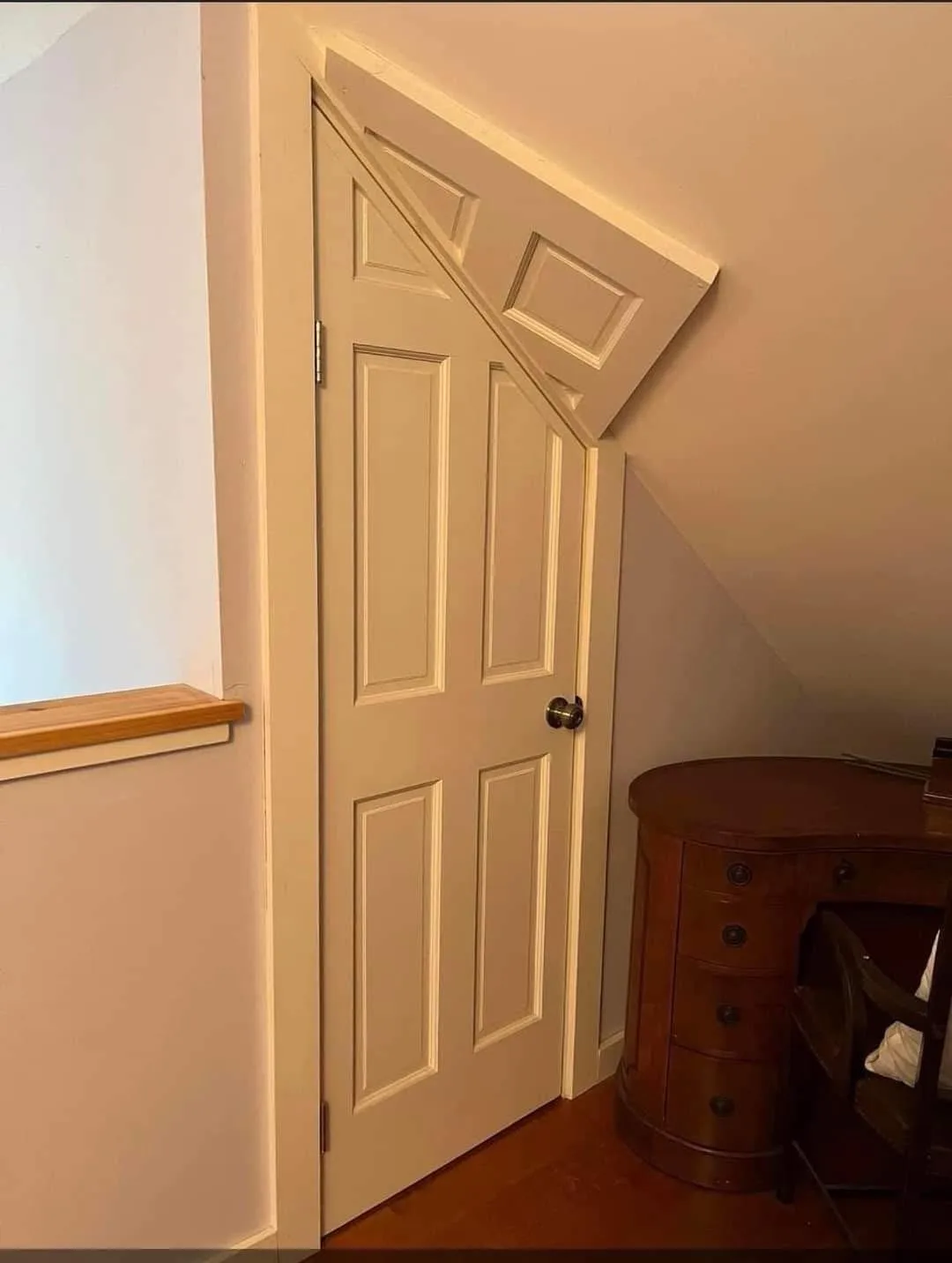 A white paneled door in a sloped attic room is visibly angled upward, wedged into the ceiling, making it impossible to open…