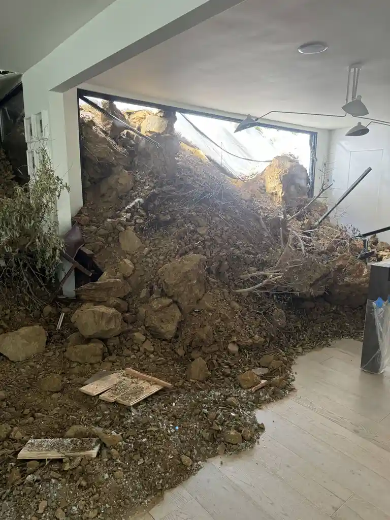 A modern interior room with light wood flooring and white walls is partially destroyed by a massive landslide of dirt…