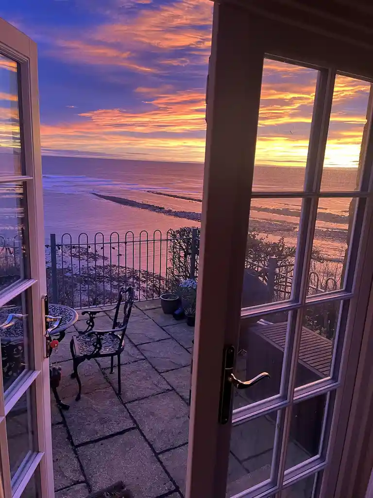 A view from inside a house looking out through open french doors onto a stone patio with a wrought iron chair and table…