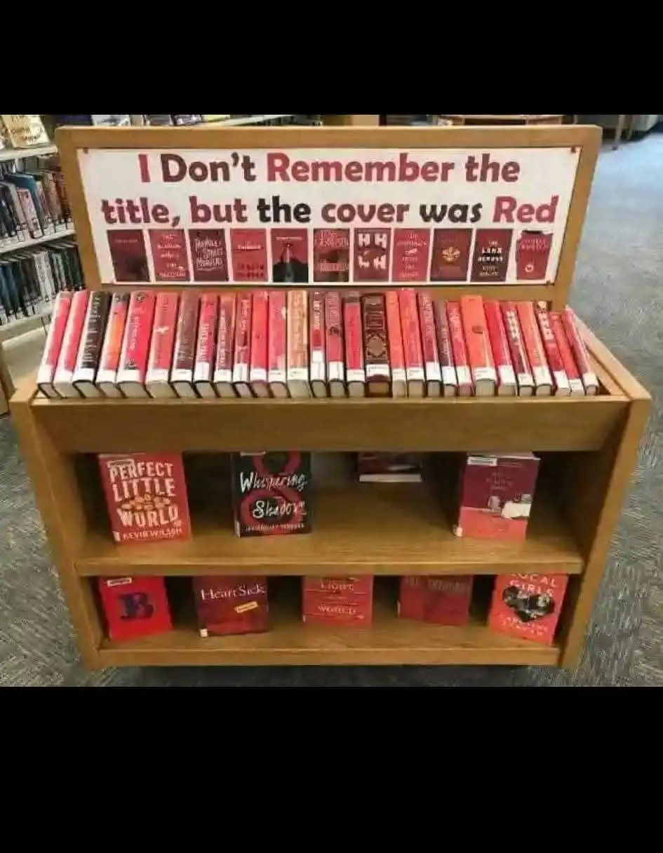 A wooden bookshelf in a library displays numerous books with red covers. The top of the shelf has a sign reading 'I Don't…
