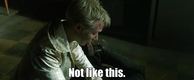 A man with blond hair, wearing a stained white shirt and dark pants, sits slumped on a checkered floor in a dimly lit room…