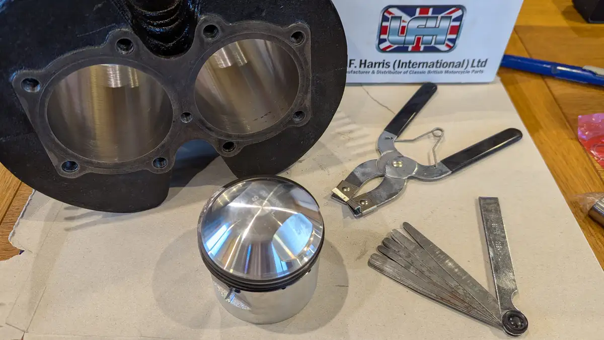 A close-up photo shows a shiny new piston sitting next to a motorcycle engine block with two cylinders, alongside tools…
