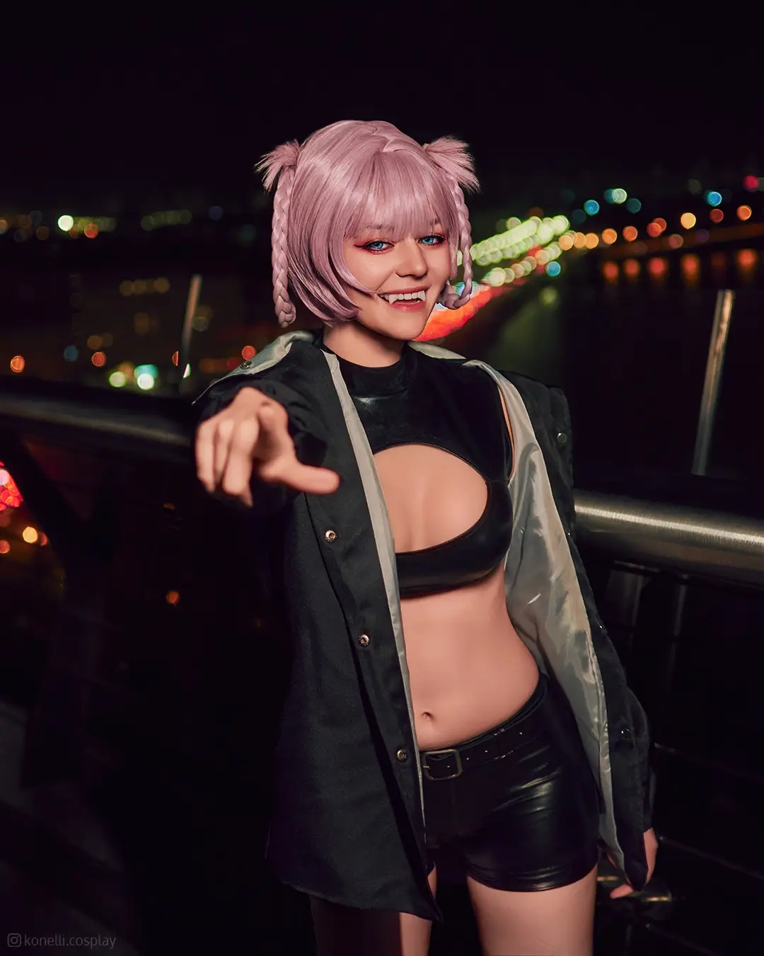 A person in a cosplay outfit with pink braided hair and cat ears points directly at the viewer with a wide, toothy grin…