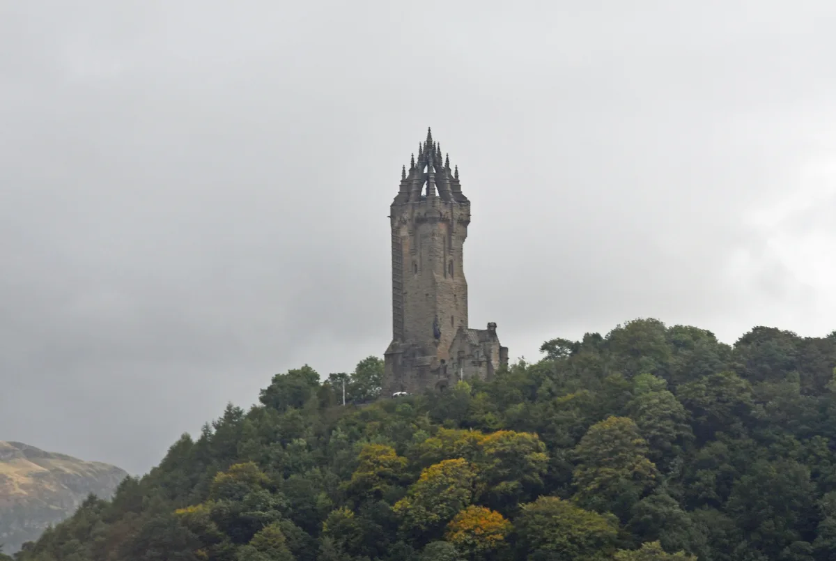 A tall, ornate stone tower with spires stands atop a densely forested hill under an overcast sky. The surrounding trees are…