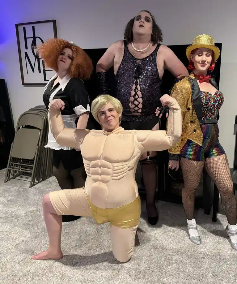 Four people in elaborate costumes pose indoors; one in a tan inflatable muscle suit kneels front and center flexing, while…