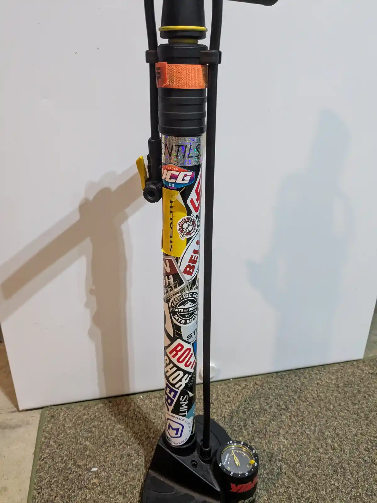 A black bicycle floor pump covered in numerous colorful stickers from bike brands and shops, standing upright against a…