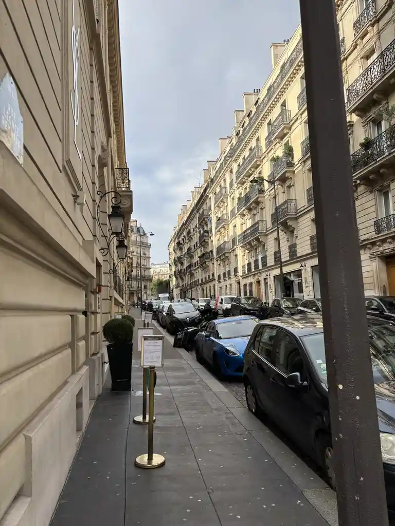 A narrow Parisian street with classic Haussmann-style buildings, parked cars including a blue sports car, and a YSL logo on…