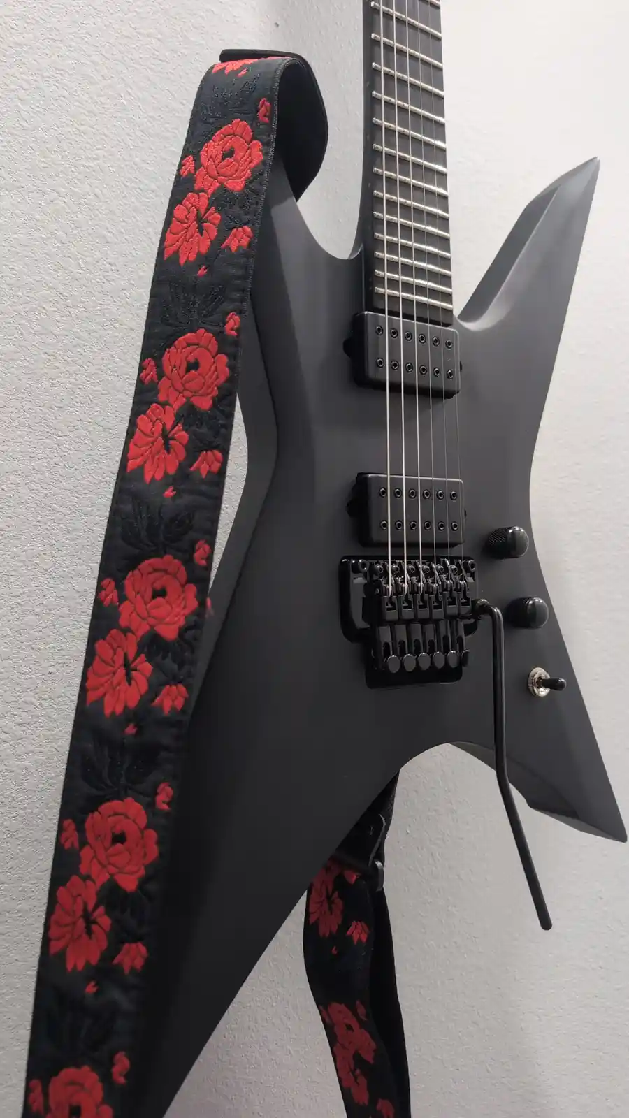 A black electric guitar with a sharp, angular body and black hardware is mounted on a white wall. A black guitar strap with…