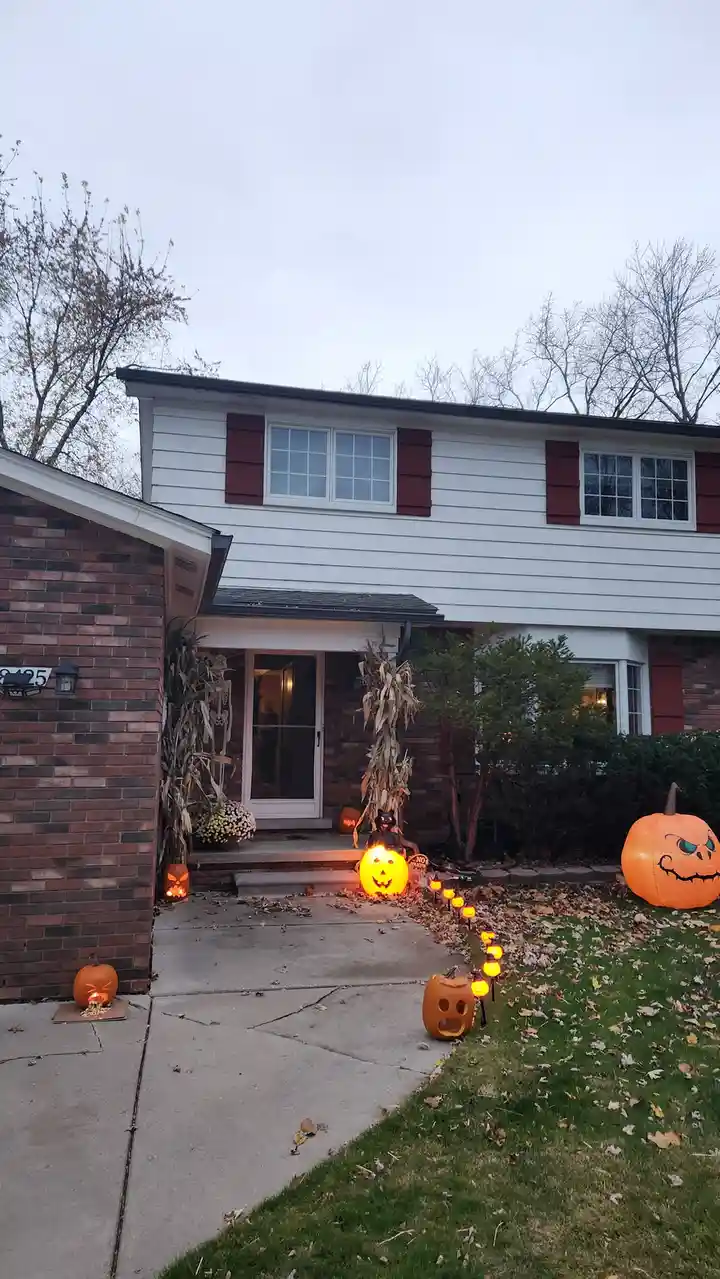 A house decorated for Halloween with numerous glowing jack-o'-lanterns, an inflatable pumpkin, cornstalks, and a lit…