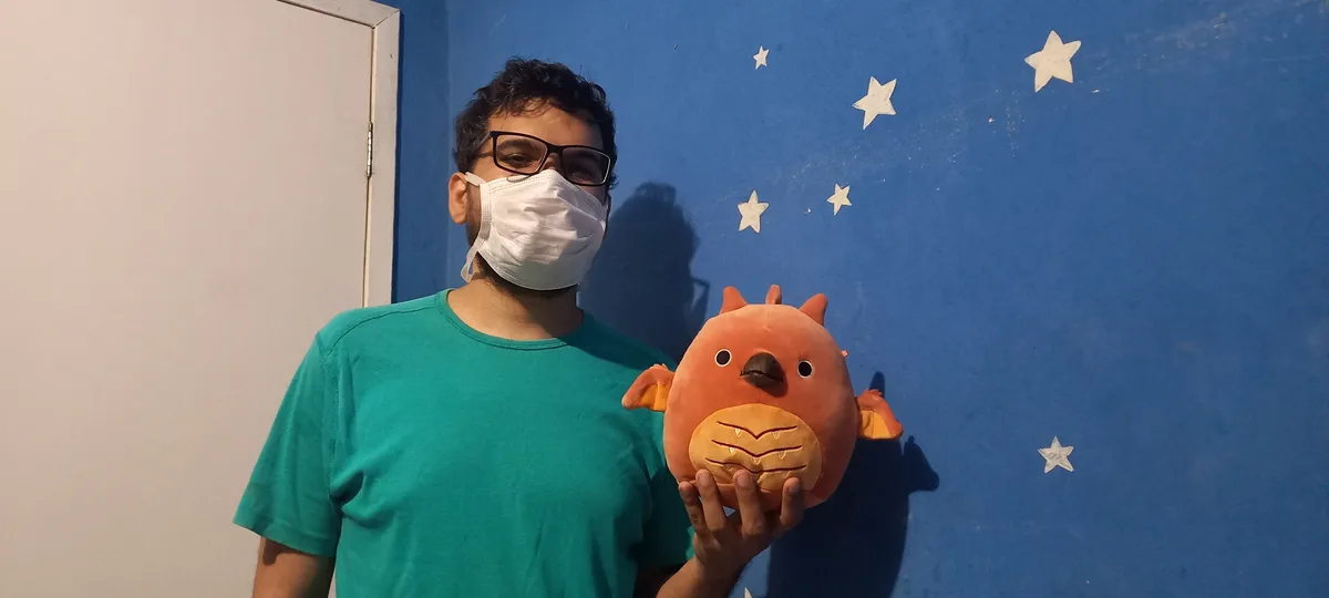 A man wearing glasses and a white face mask stands in front of a blue wall with white stars, holding an orange plush toy…