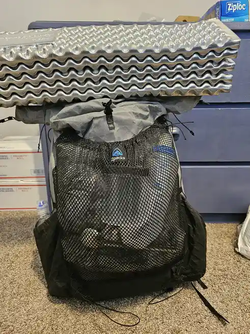 A black mesh 60L Zpacks backpack sits on a carpeted floor, with a stack of four silver, wavy, ridged foam camping pads…
