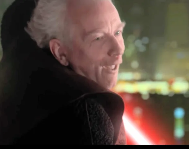 a close-up of a man with white hair and a dark robe, smiling broadly while looking off-camera, with a red lightsaber visible…