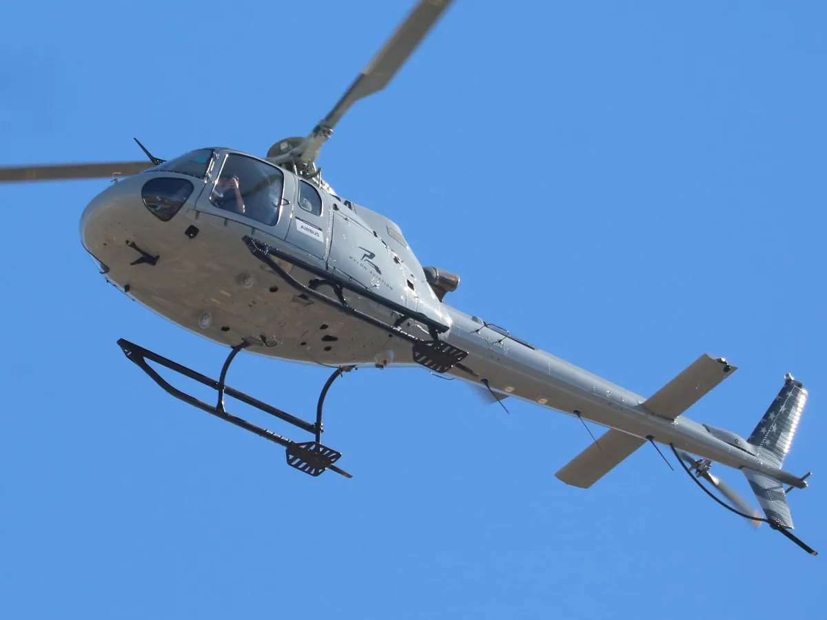 A gray Airbus helicopter is flying against a clear blue sky, with a person visible inside the cockpit holding up a phone…