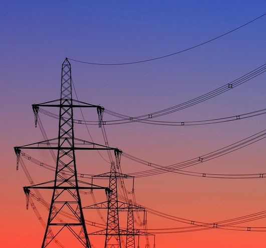 A silhouette of multiple electricity pylons against a gradient sunset sky transitioning from orange to purple. Overlaid text…