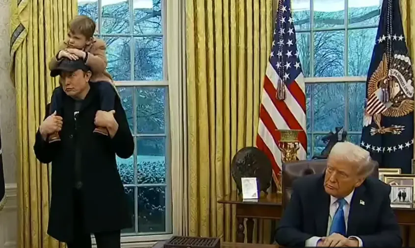 A man in a black coat carries a small child on his shoulders while standing in the Oval Office, facing a seated Donald Trump…