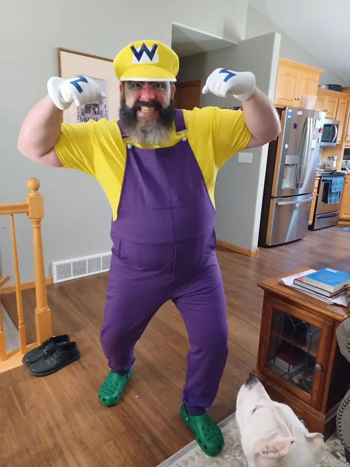 A man with a beard and blue eye makeup poses in a Wario costume—yellow shirt, purple overalls, yellow cap with a W, and…