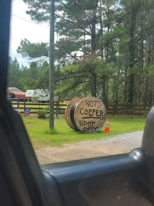 A large wooden spool sits on a grassy area beside a gravel road, with handwritten text on its side reading 'NOT COPPER Fiber…