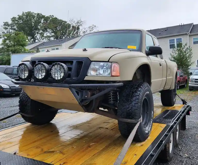 A heavily modified, tan Ford F-150 pickup truck with oversized tires, a custom front bumper, and three large KC lights is…