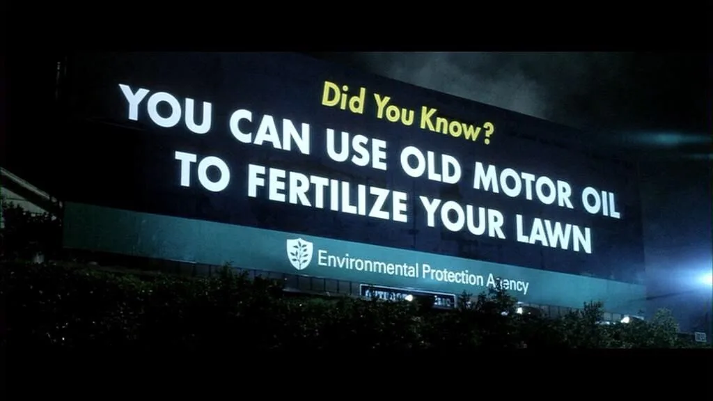 A nighttime shot of a large illuminated billboard that reads 'Did You Know? YOU CAN USE OLD MOTOR OIL TO FERTILIZE YOUR…