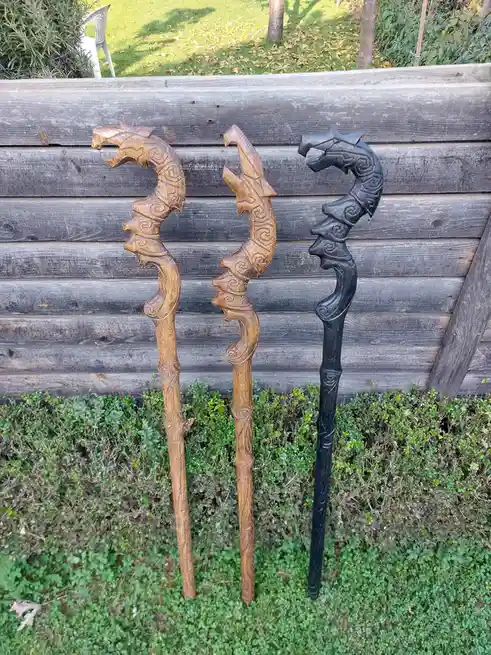 Three intricately carved wooden staffs, each topped with a dragon head, stand upright in a grassy area against a weathered…