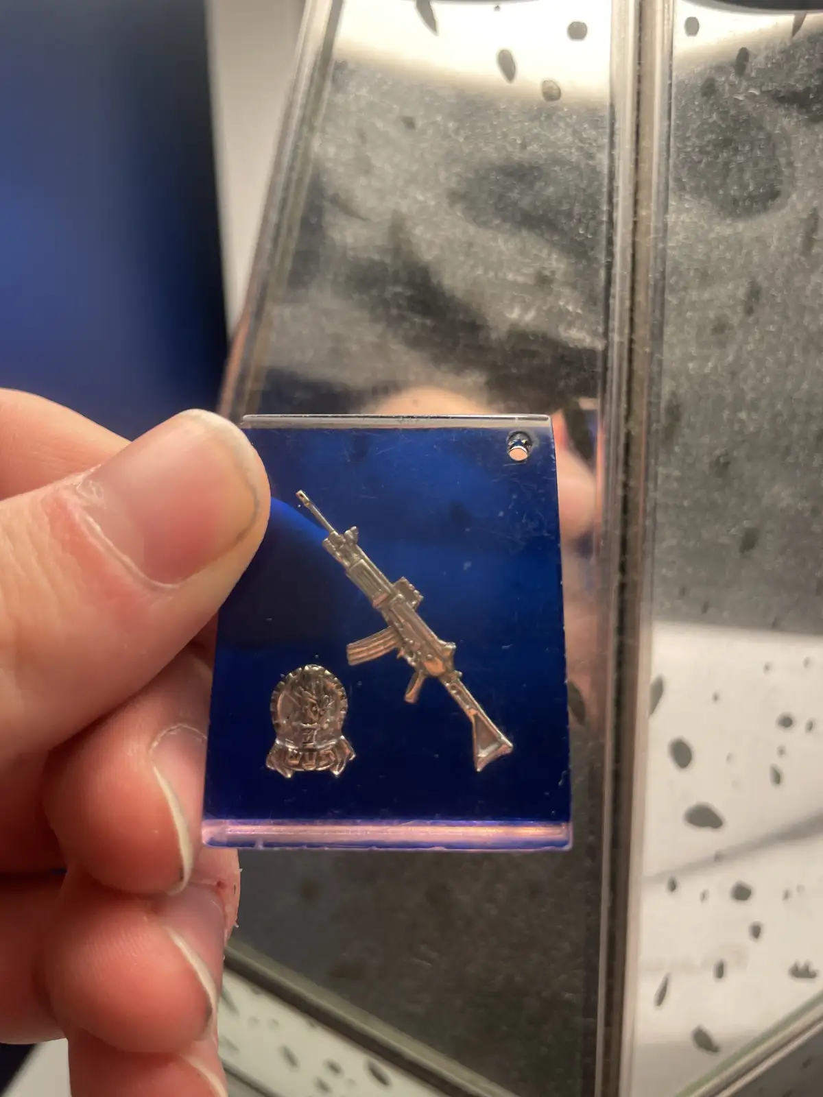 A hand holds a small blue acrylic pin displaying a miniature silver rifle and a circular police badge emblem. The background…