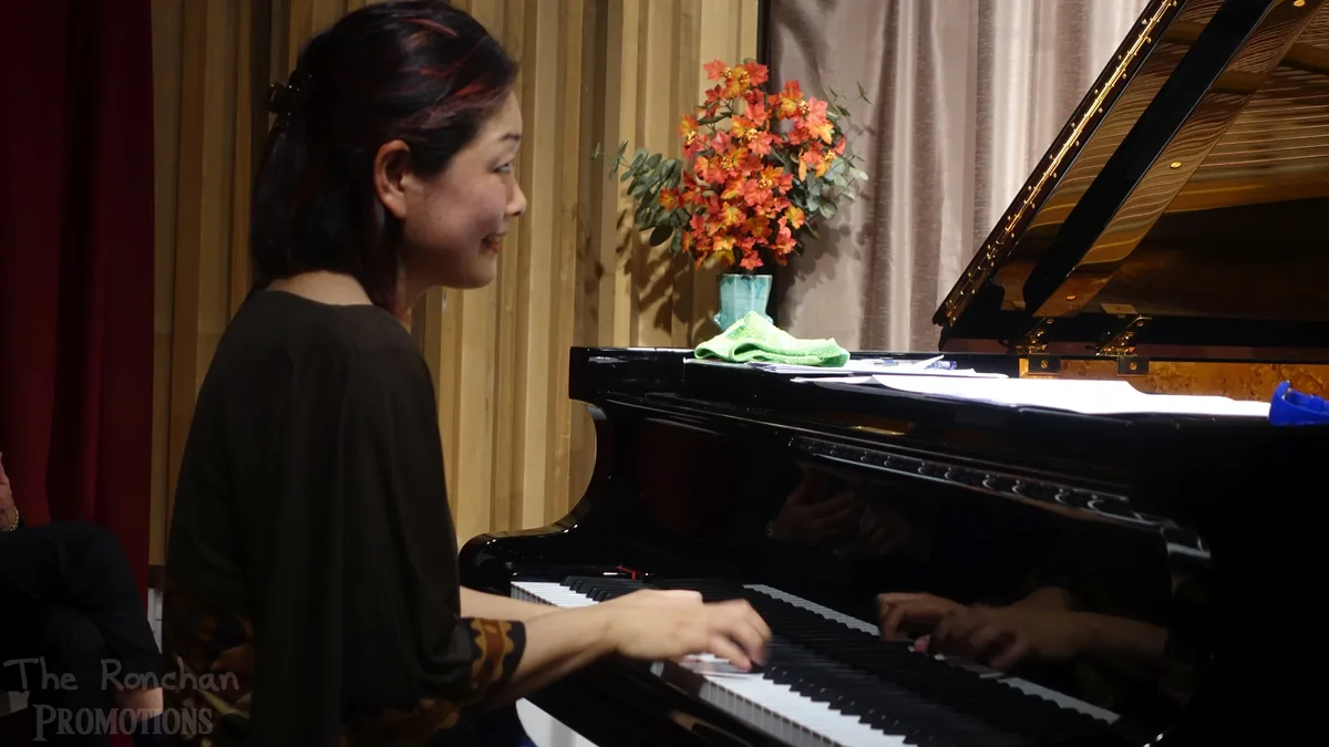 A woman with dark hair and red streaks smiles while playing a black grand piano. Sheet music and a green cloth rest on the…