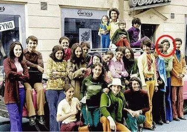 a vintage group photo of many people posing together on a street, with a red circle highlighting a man standing slightly…