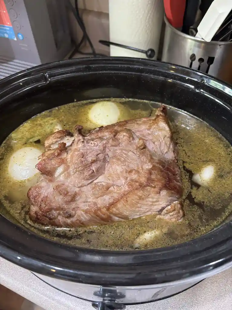 A close-up photo shows a large, seared piece of meat, likely beef, submerged in a yellowish broth with whole onions inside a…