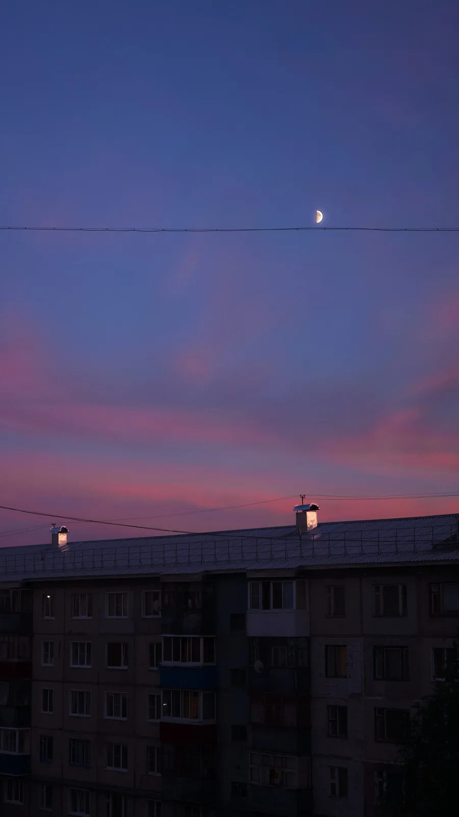 A serene twilight photo shows a crescent moon positioned directly above a thin power line strung across the sky, with a…