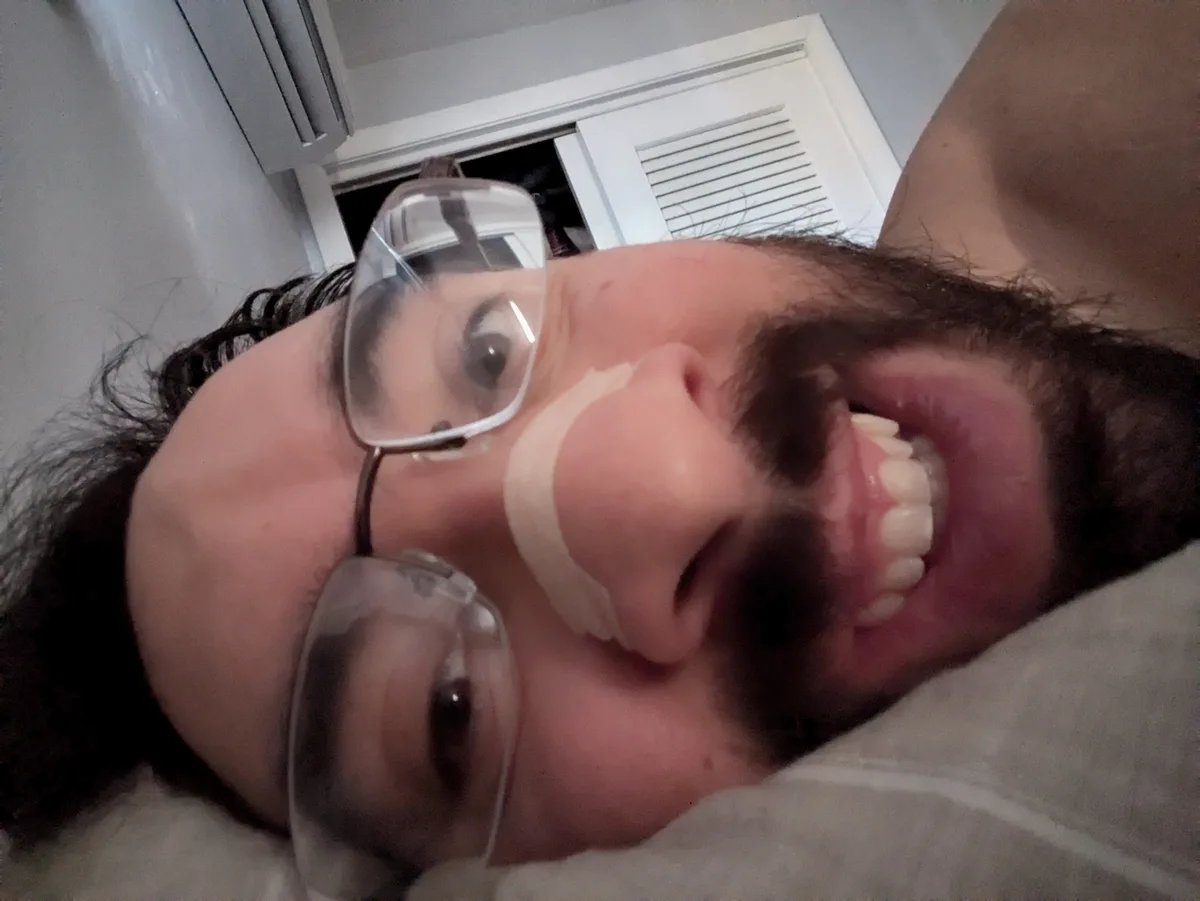 A person with glasses and a beard lies on their back, looking up at the camera with a wide, exaggerated smile. A strip of…