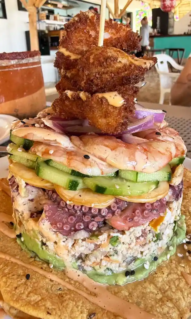 a towering, layered seafood dish sits on a tortilla base, featuring avocado, octopus, cucumber, shrimp, and fried pieces on…