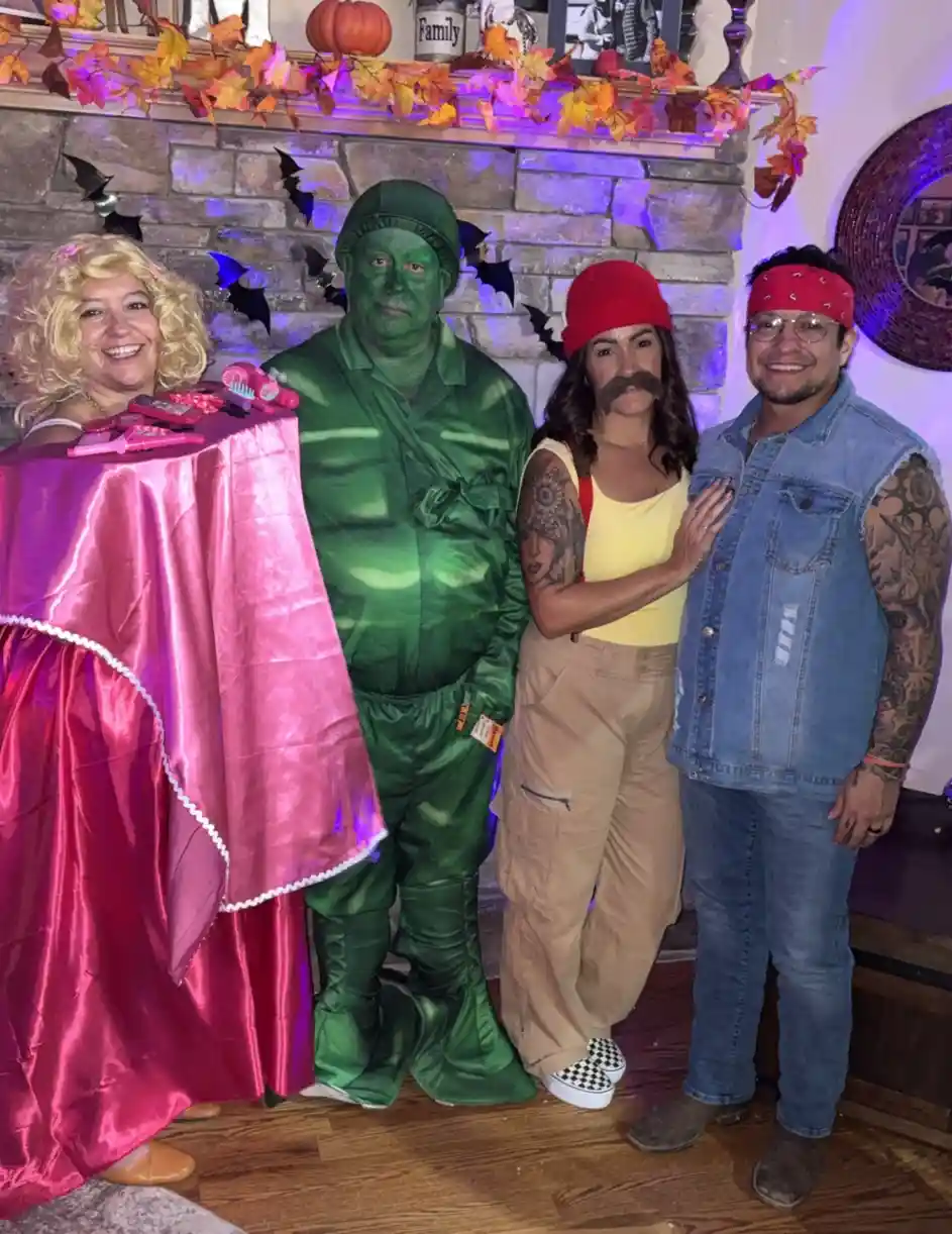 Four people pose together in a Halloween setting with autumn decorations and bats on a stone fireplace. From left to right…