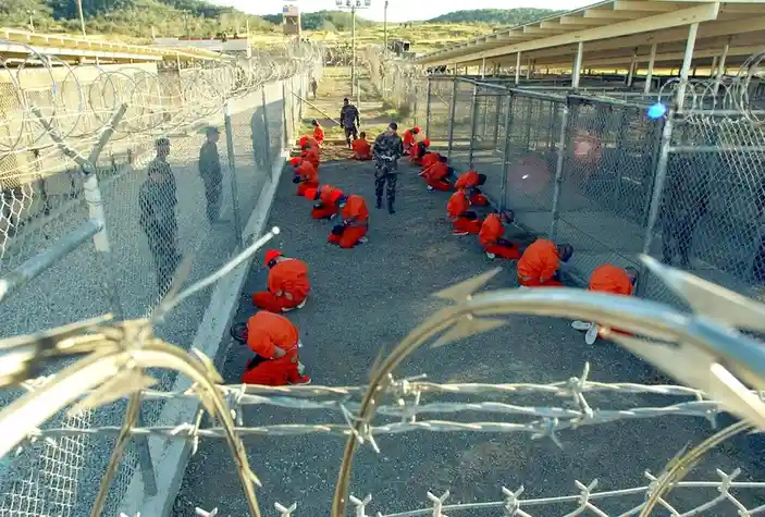 A group of people in orange jumpsuits are kneeling in a line inside a high-security prison yard, surrounded by chain-link…
