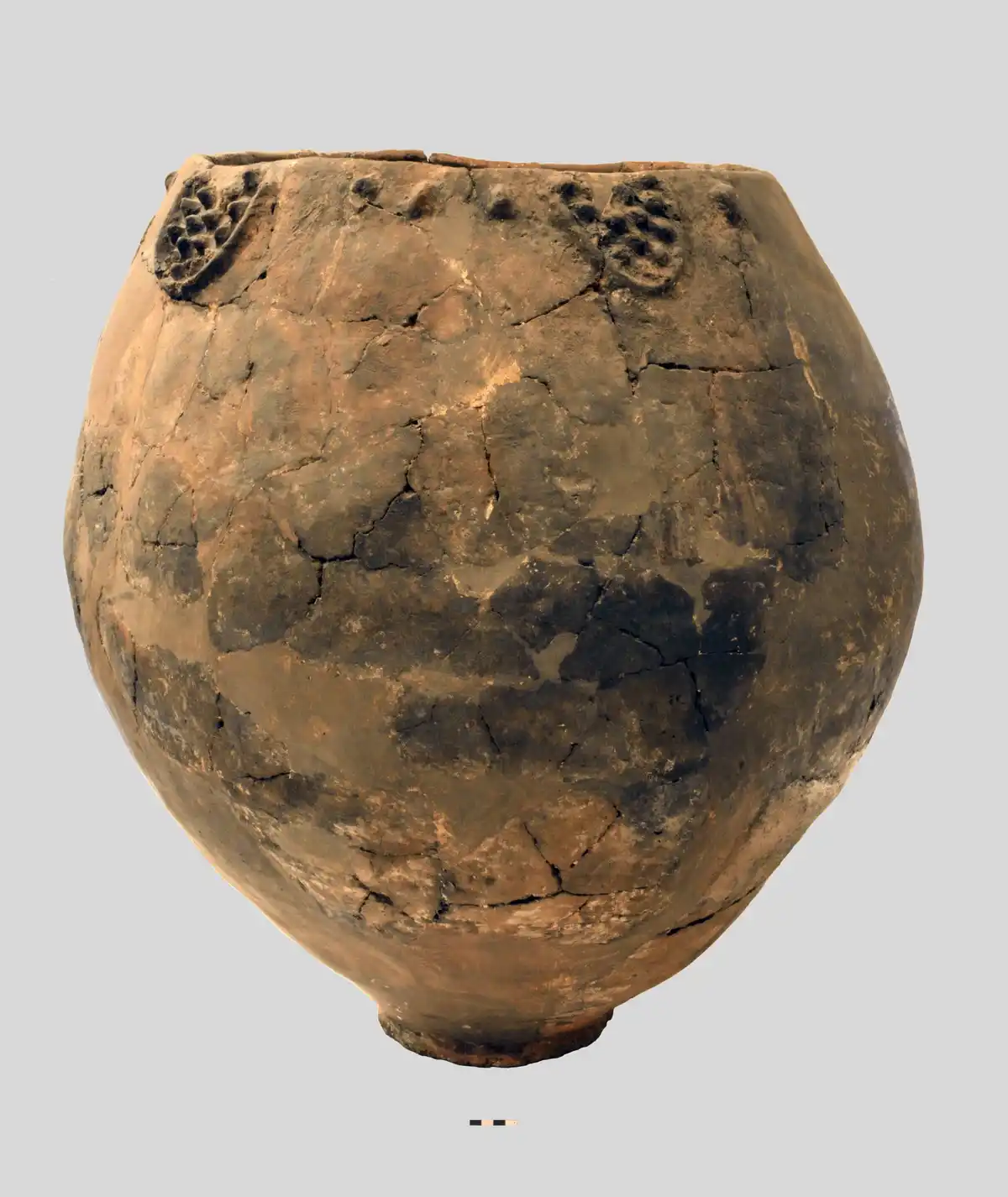 The image shows a large, round, ancient clay pot with visible cracks and patches of restoration, featuring decorative…