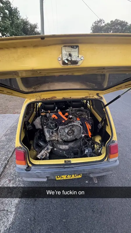 A yellow car with its trunk open, revealing a disassembled engine and mechanical parts inside. The overlaid text reads…