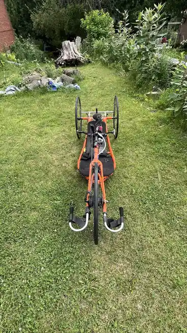 An orange recumbent trike is parked on a grassy lawn, facing away from the camera, with a tree stump and logs visible in the…