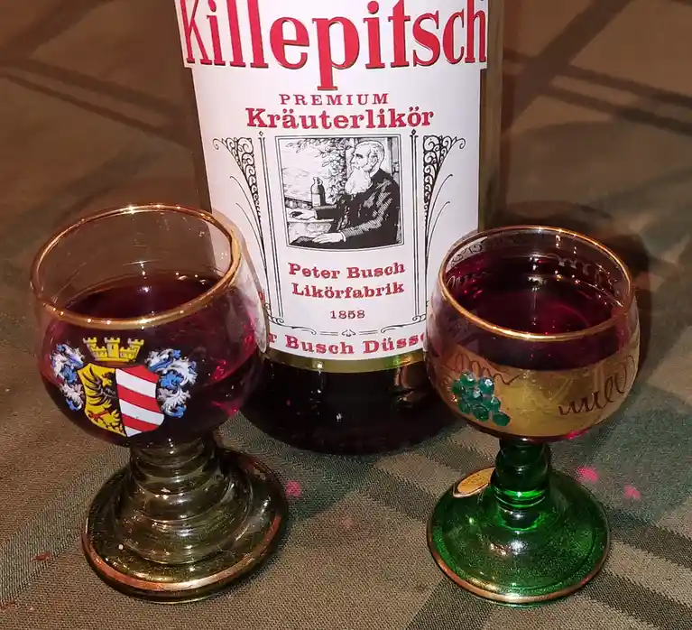 A bottle of Killepitsch Premium Kräuterlikör by Peter Busch Likörfabrik from 1858 stands between two ornate glasses filled…