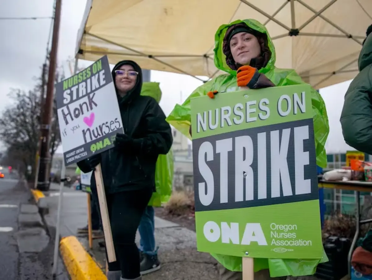 Two women in rain gear stand outdoors under a canopy holding protest signs. The woman in the foreground wears a bright green…