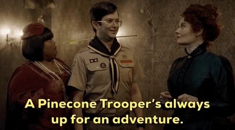 Three people in a dimly lit, rustic room. A man in a Boy Scout uniform with glasses stands between a woman in a red outfit…