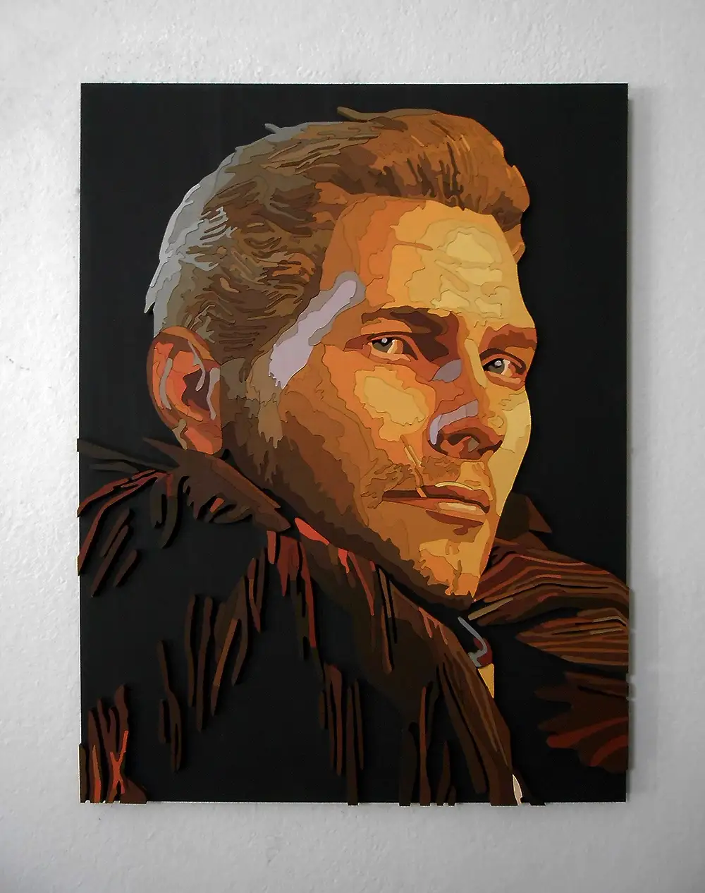 A stylized, layered papercut portrait of a man with a serious expression, mounted on a white wall. The artwork uses warm…