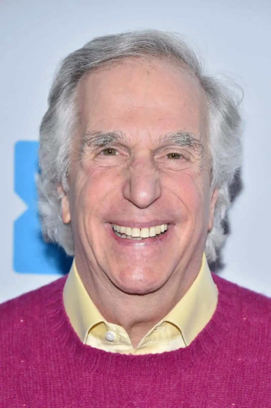 A close-up photo of an older man with gray hair, wearing a magenta sweater over a yellow collared shirt, smiling broadly…