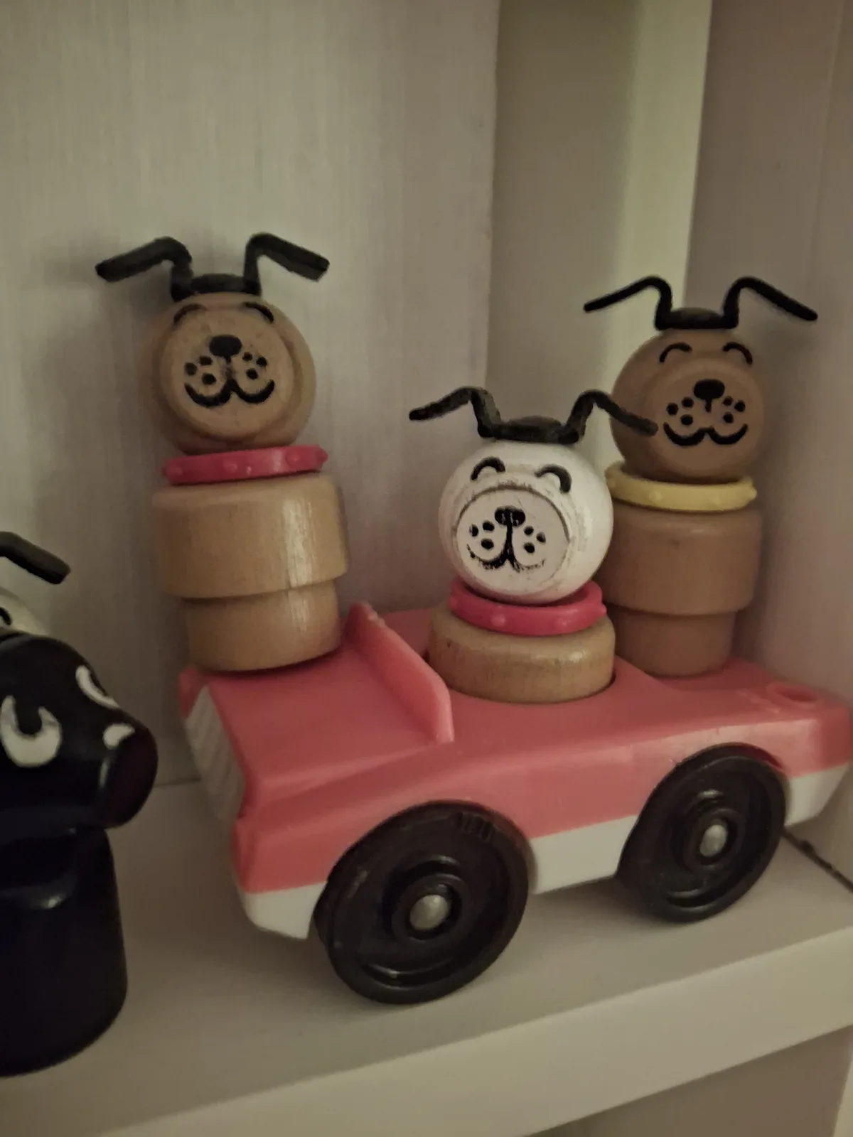 Three wooden dog figurines with painted faces and collars are stacked on a pink toy car; one white dog in the center has a…