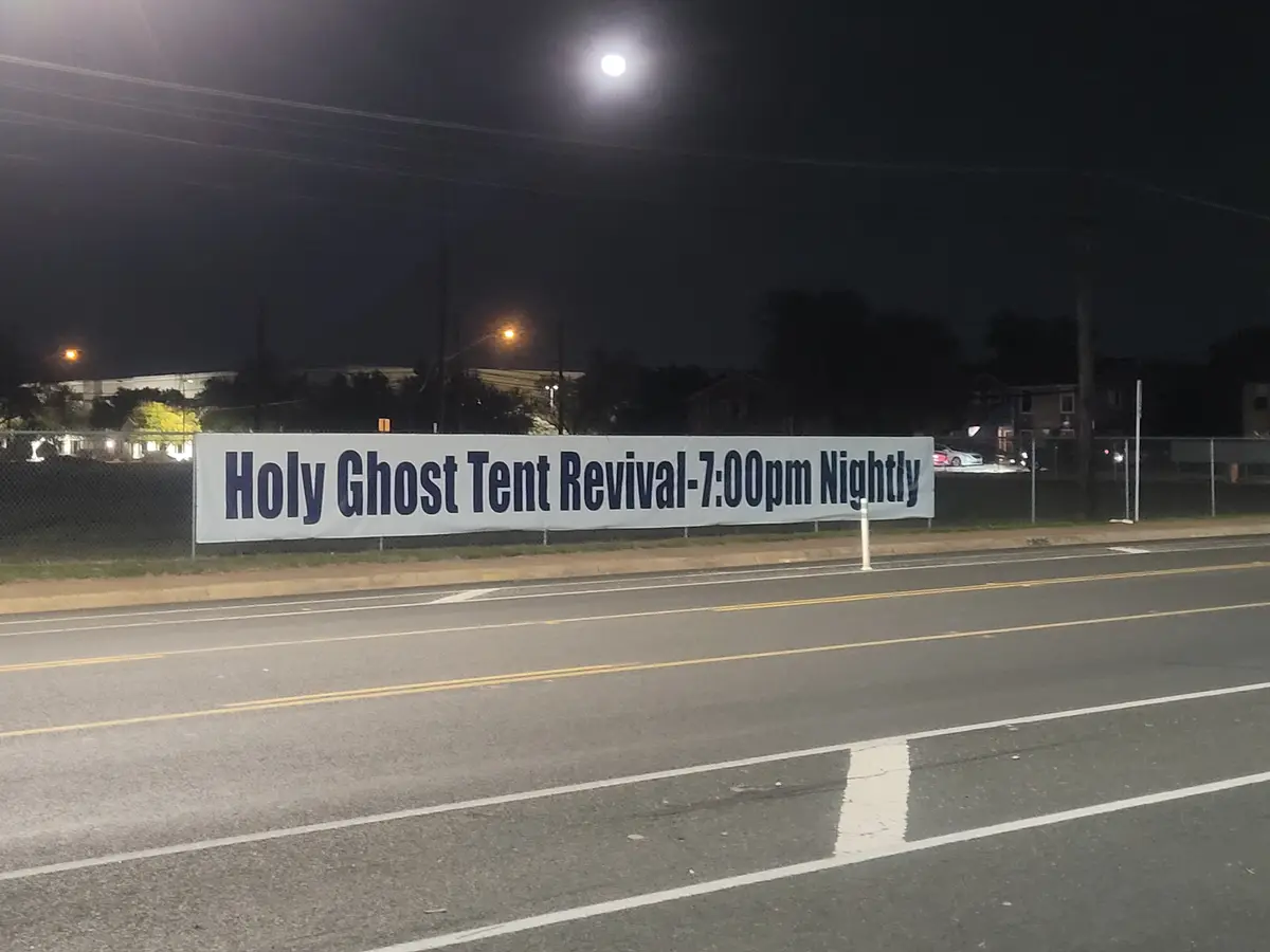 A nighttime photo shows a large white banner attached to a fence beside a road, under a bright full moon. The banner reads…