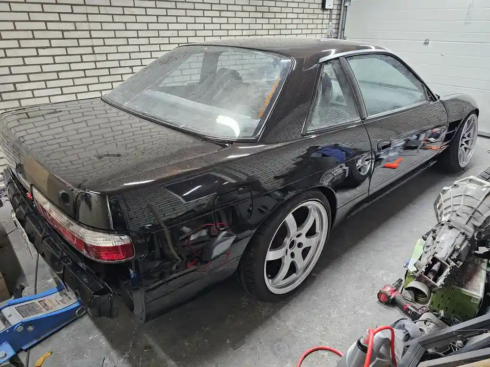 A glossy black Nissan Skyline R32 is shown in a garage with its rear bumper removed and a blue jack underneath, surrounded…