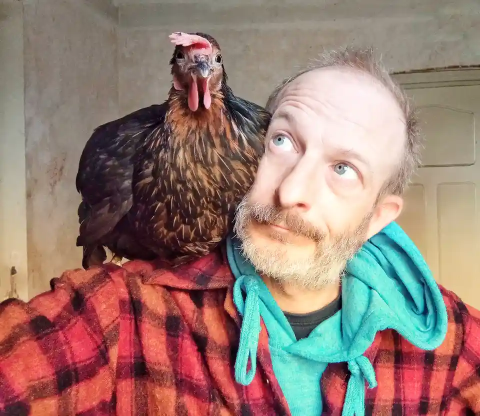 A man with a beard and blue eyes, wearing a red plaid shirt over a teal hoodie, looks upward with a slightly amused…