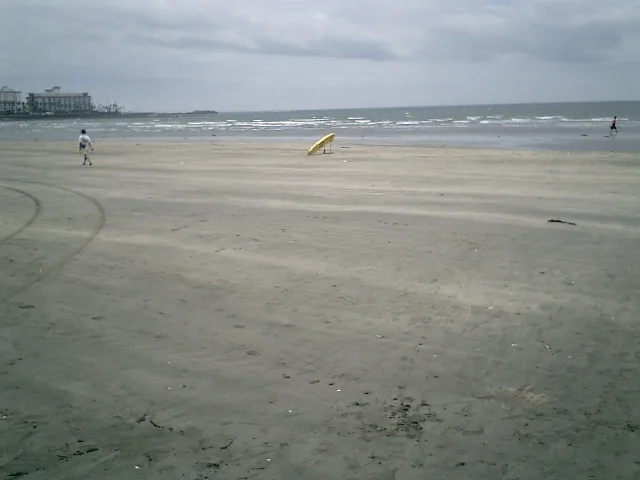 A wide, empty beach under an overcast sky with a lone yellow surfboard standing upright in the sand. Two people are visible…