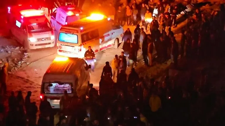 A nighttime scene shows multiple ambulances with flashing lights parked on a dirt road surrounded by a large crowd of…