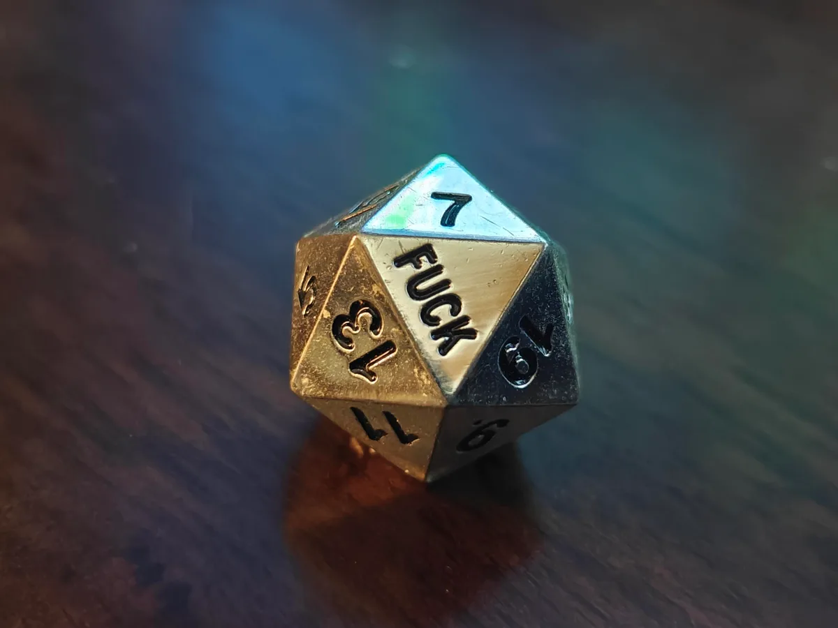 A close-up of a metallic, multi-colored 20-sided die resting on a dark wooden surface, showing the number 7 on top and the…