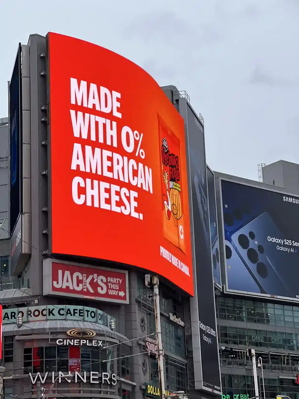 A large red billboard in a city displays the text 'MADE WITH 0% AMERICAN CHEESE.' and 'PROUDLY MADE IN CANADA.' alongside an…