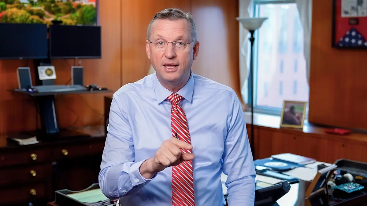 a man in a light blue shirt and red striped tie stands in an office, pointing directly at the viewer with a serious…